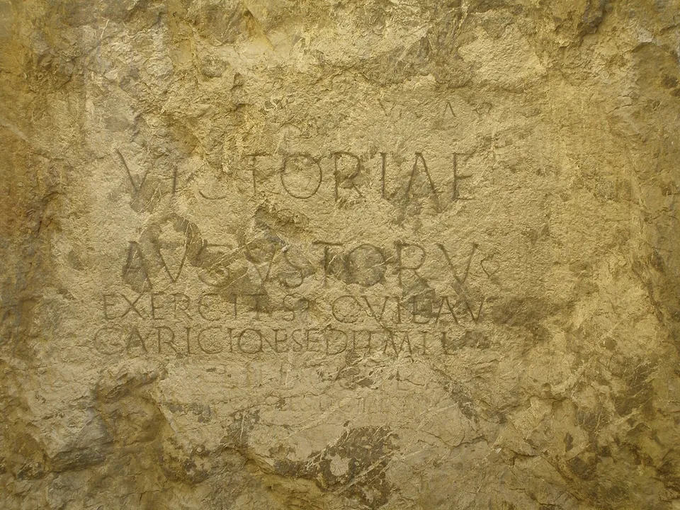 A close view of carved Roman capitals on weathered yellowish rock, the words VICTORIAE and AVGVSTORV large and clear, two smaller lines below them and fainter traces of further lines at the bottom.