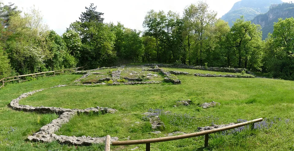 Low rubble wall footings running across a spring meadow on a wooded hilltop, a wooden rail in front and rocky cliffs rising behind the trees.