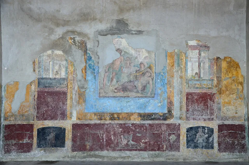 A plastered wall painted in panels of deep red, blue and ochre, with painted architecture at either side and, at the centre, a figure scene of a reclining woman with other figures around her, the plaster broken away in patches.