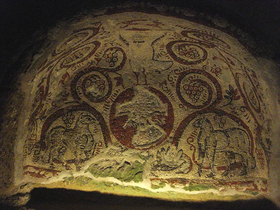 A mosaic filling a low arched niche, lit from below. Red-outlined vine tendrils curl into roundels holding bunches of grapes, a four-legged animal, probably a lamb, stands in each lower corner, and the central roundel is largely lost.