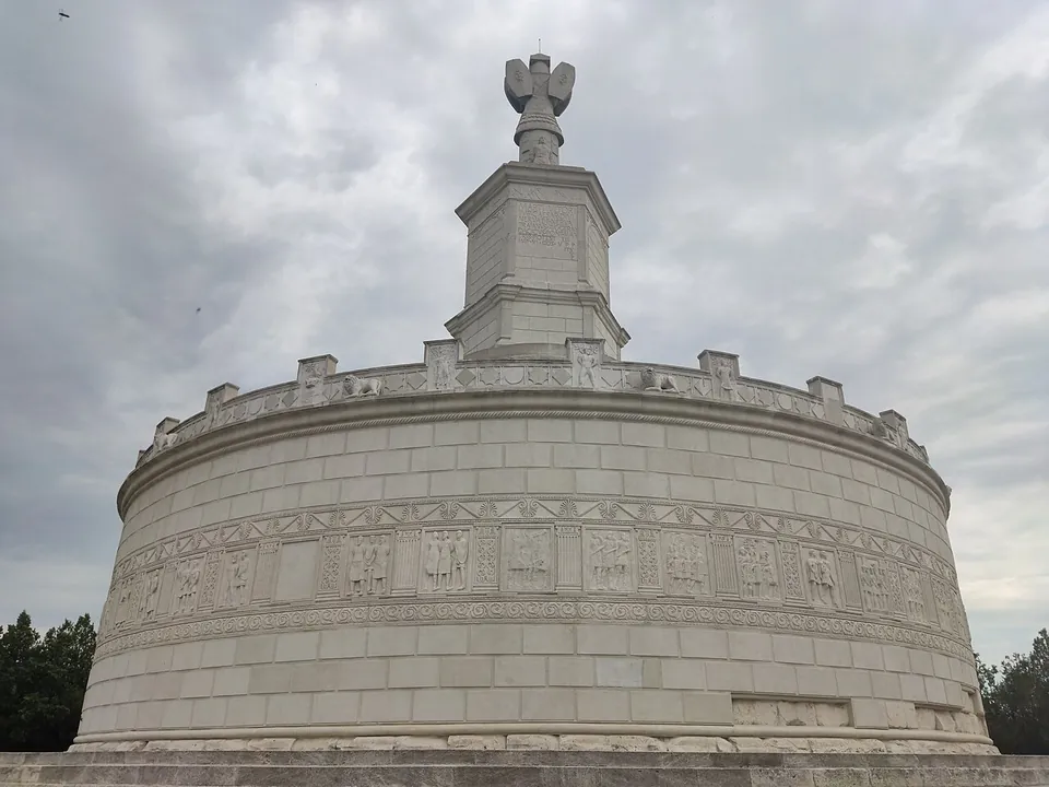 The white stone drum of the monument seen from close below, ringed by a band of square relief panels with figures between decorated friezes, under a crenellated parapet and the central tower.