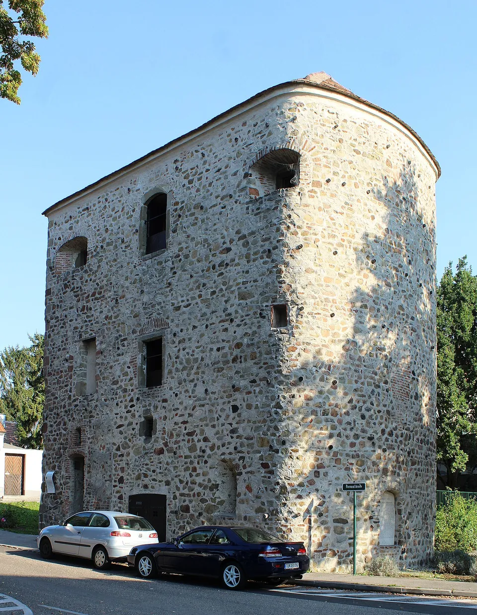 A tall, roughly square stone tower with a rounded corner and small irregular windows, its walls of rubble masonry, standing beside a street with parked cars.
