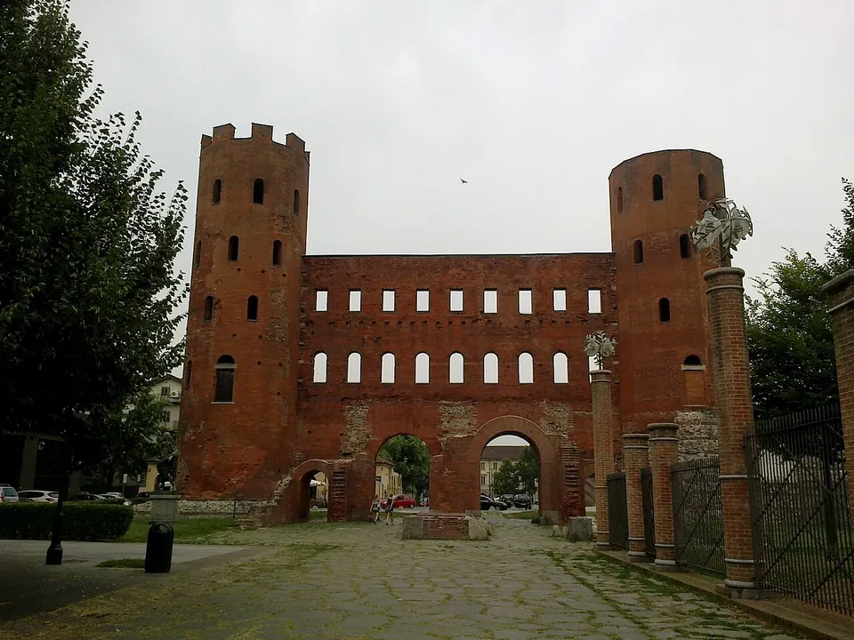The brick gate front-on at the end of a stone-paved street, with arched openings at ground level, a row of arched windows and a row of square windows above, between two battlemented towers; a bronze statue stands on a tall brick pedestal to the right.