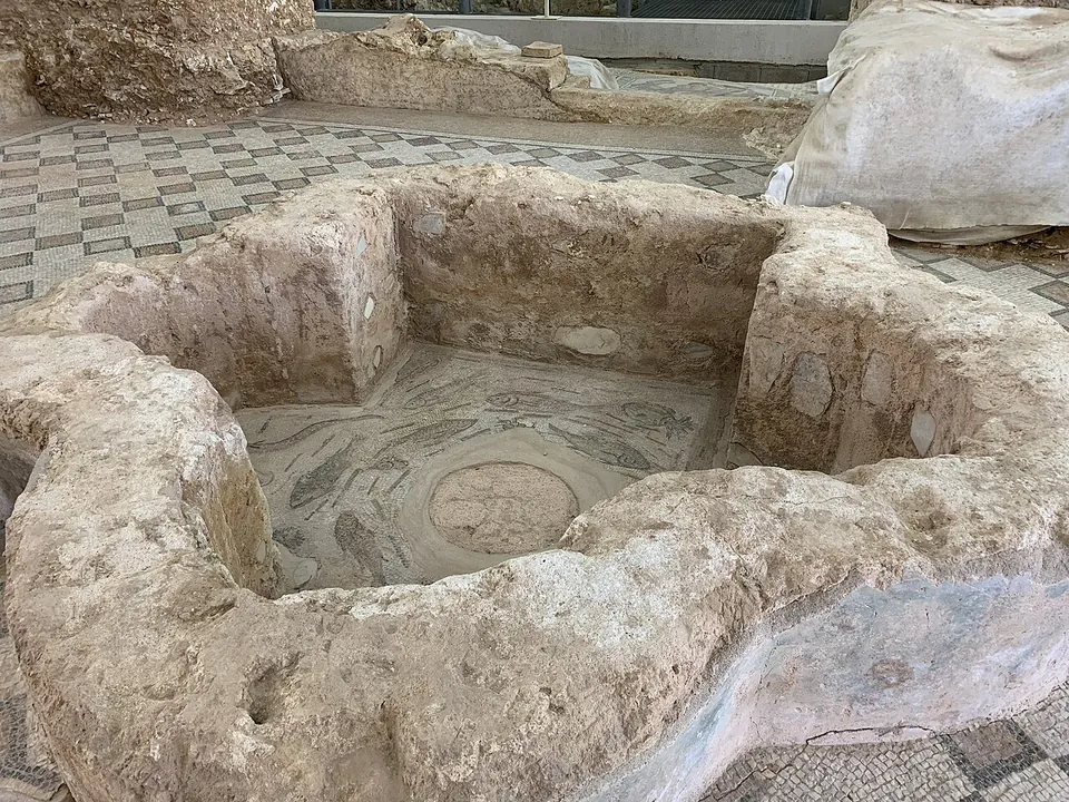 A low basin of rough stone with a mosaic on its floor, set into a pavement of chequered mosaic, with a protective sheet over the ruins beyond.