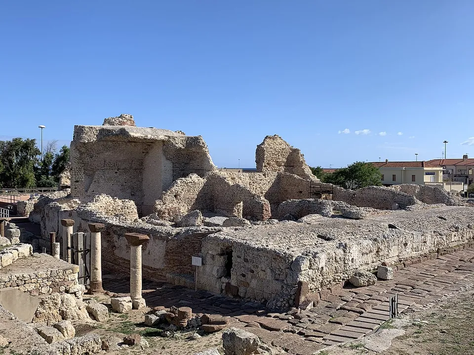 Ruined walls and low rooms of stone and brick line a street paved with large dark slabs, with a row of short columns beside it and the houses of the modern town behind.