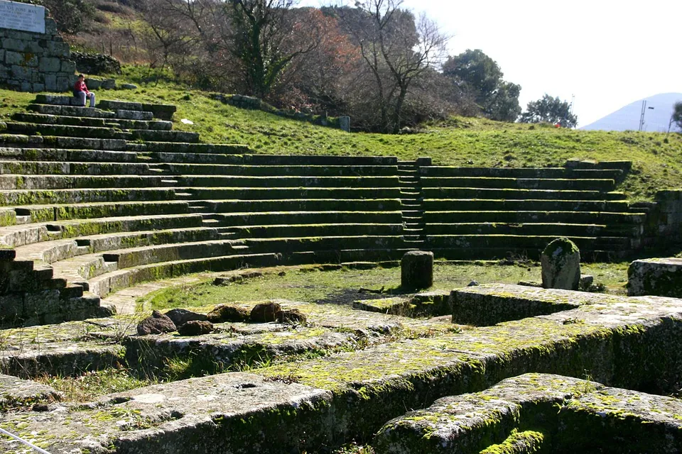 The curved stone seating of a small Roman theatre set into a grassy hillside, with low walls and blocks of the stage area in the foreground and bare trees on the slope above.