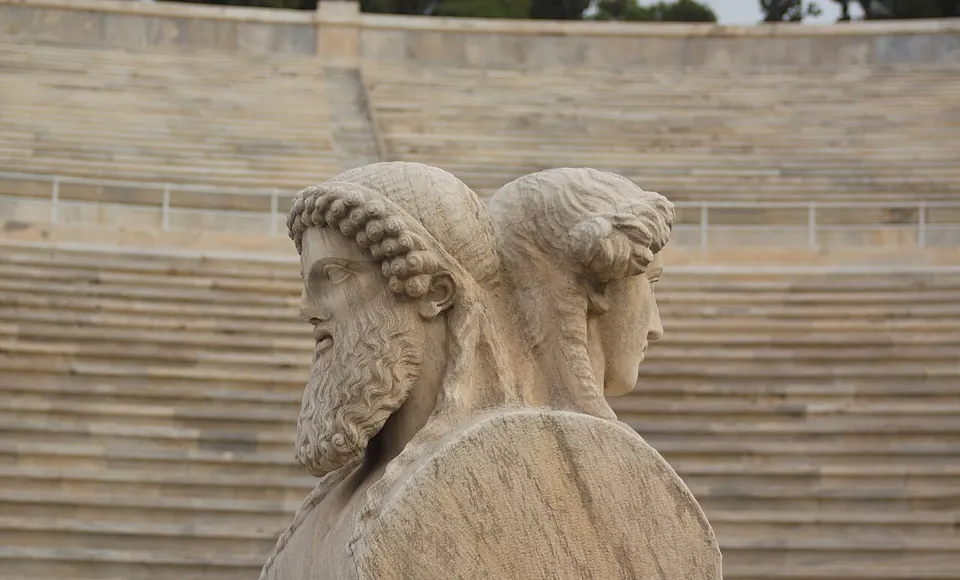 A marble pillar topped by two heads back to back, a bearded man with curled hair and a younger beardless face, with the stadium's marble seats blurred behind.