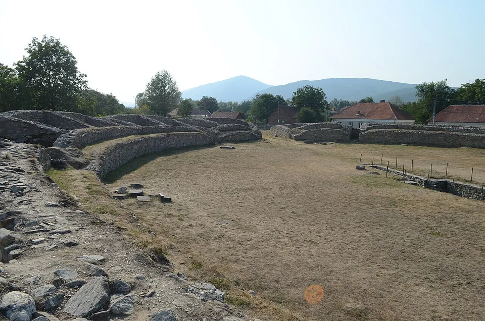 The oval arena of an amphitheatre, a flat patch of dry grass enclosed by low curving stone walls, with trees, village roofs and a hill beyond.