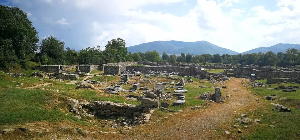 A wide view across excavated stone foundations, fallen blocks and a few standing column stumps in rough grass, with a dirt path, trees and wooded mountains under a cloudy sky.