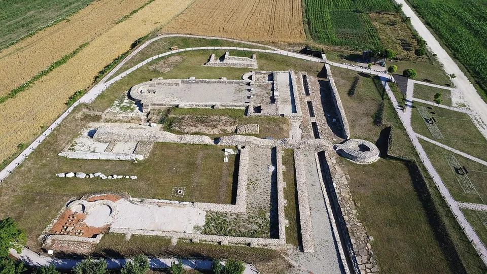An aerial view of a long rectangular hall ending in a semicircle, a round building beside it and the walls of further excavated buildings, framed by gravel paths and fields.