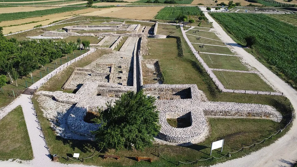 An aerial view over excavated stone walls and foundations laid out in mown grass, with a gateway between two rounded towers in the foreground, a gravel path around the site and farmland beyond.