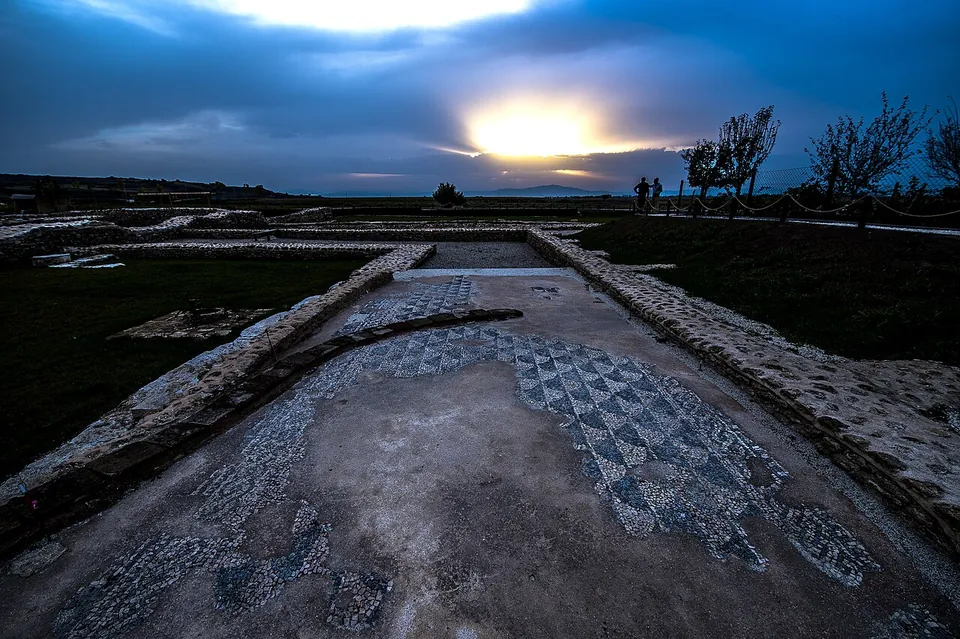 A floor mosaic of small dark and pale stones in a geometric pattern of squares and diamonds, between two low stone walls, under a dusk sky.