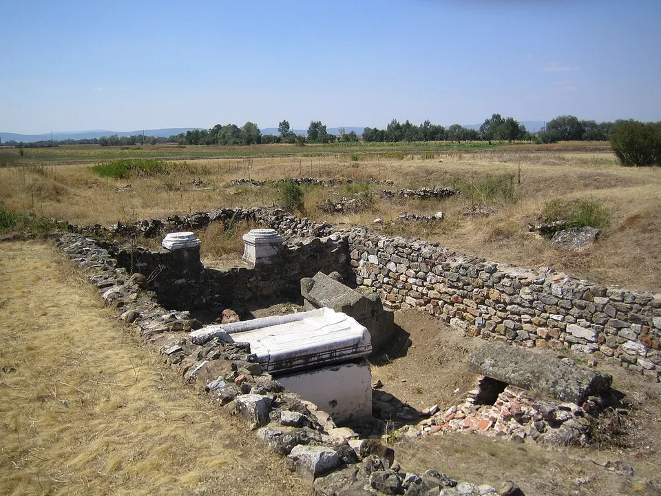 A sunken rectangle of low rubble-stone walls in dry grass, with blocks of white stone standing inside it and flat countryside and a line of trees beyond.