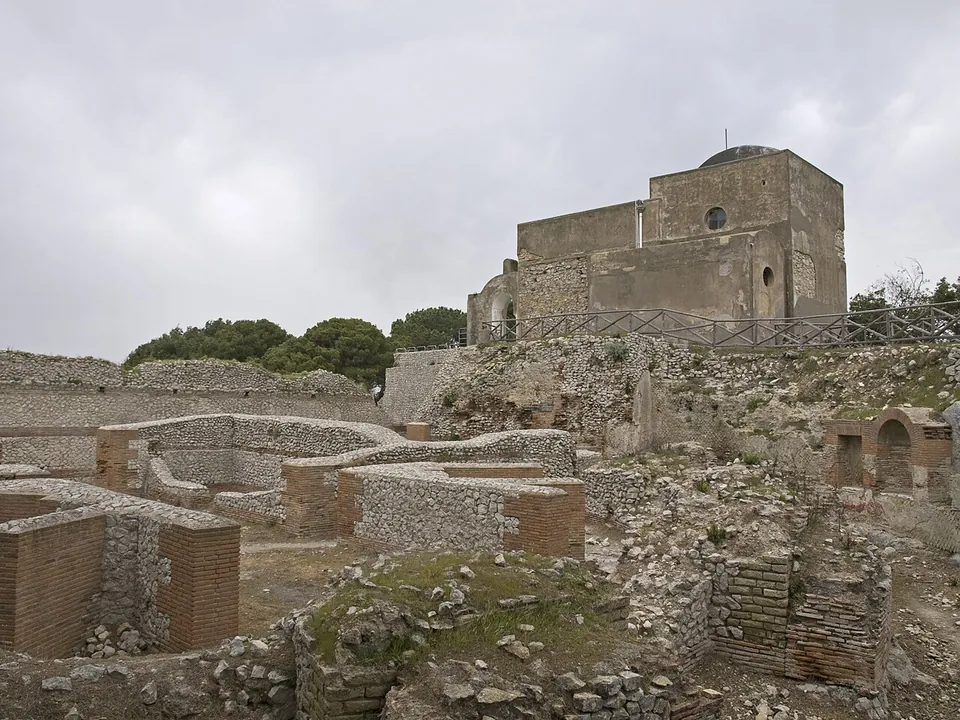 Roofless rooms of brick and rubble walls, a few metres high, fill the foreground on a hilltop under a grey sky; behind them, on higher ground, stands a plain square church with a small dome.