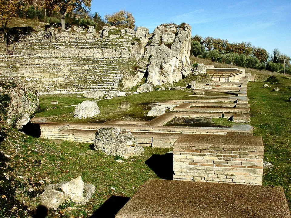 Tiers of stone seating rising against a hillside, with tall eroded masses of masonry at one end, and in the foreground the low walls of the stage buildings in the grass.