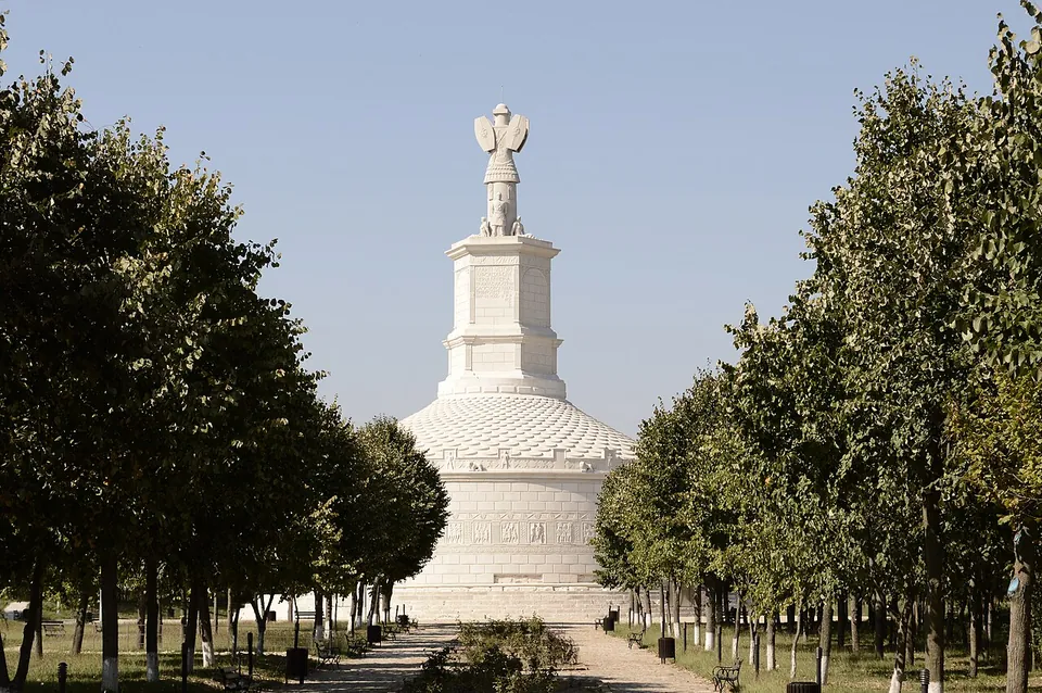 A tall white cylindrical monument with a stepped conical roof and a hexagonal tower topped by a sculpted trophy, seen at the end of a paved avenue lined with young trees.