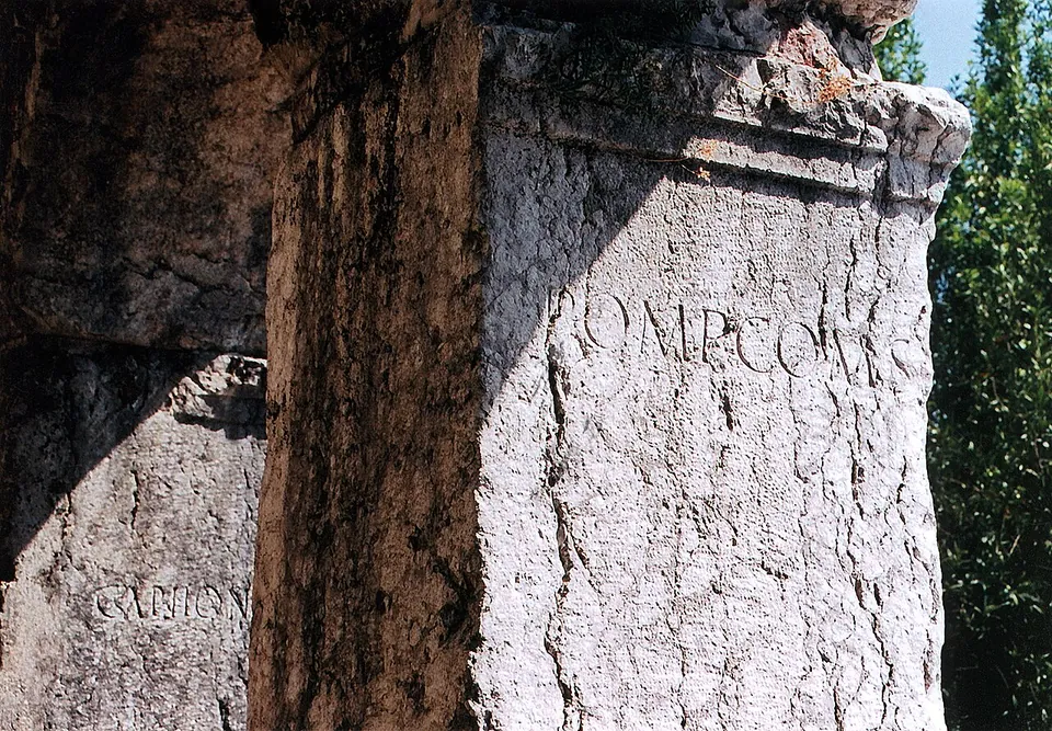 Close view of a pale stone block with large, worn Roman capital letters cut into its face, beside a shadowed block carrying more letters.