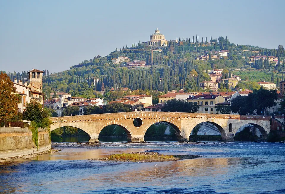 The whole bridge side-on across a wide, calm river in evening light, five round arches with a round opening above the pier between the second and third, a square tower at the left end, and a wooded hill crowned by a domed church behind.