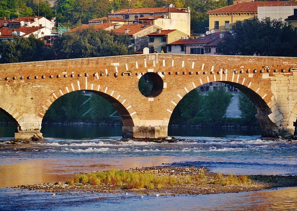 A close view of two round arches of red brick whose arch rings are edged with pale stone blocks, a round opening framed in stone in the pier between them, rippling water below and a gravel bar with grass in the foreground.