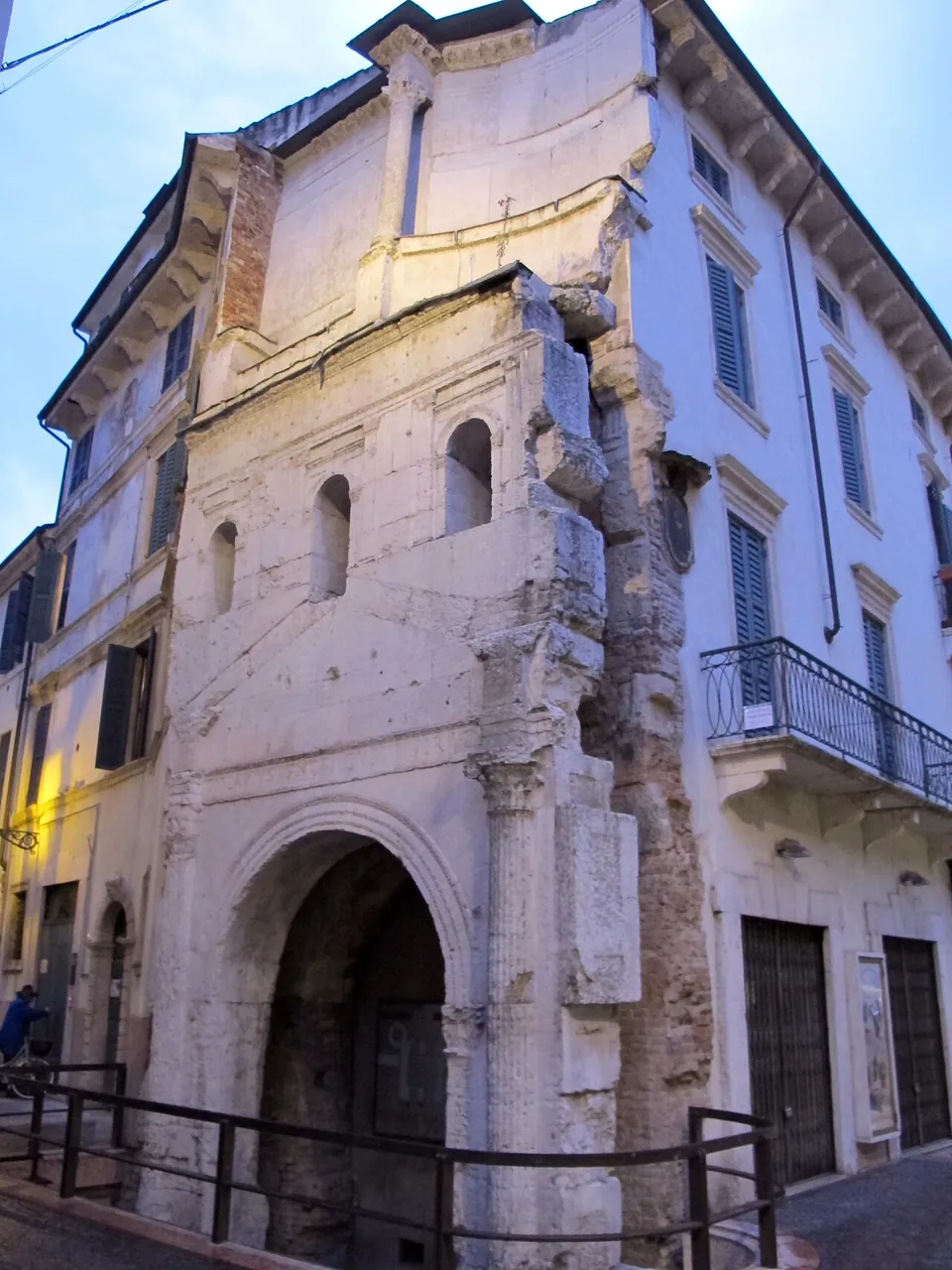A fragment of a pale stone gate front attached to the corner of a house, with one round arch at street level, a column and pilaster beside it and small arched windows above; a railing runs in front of it.