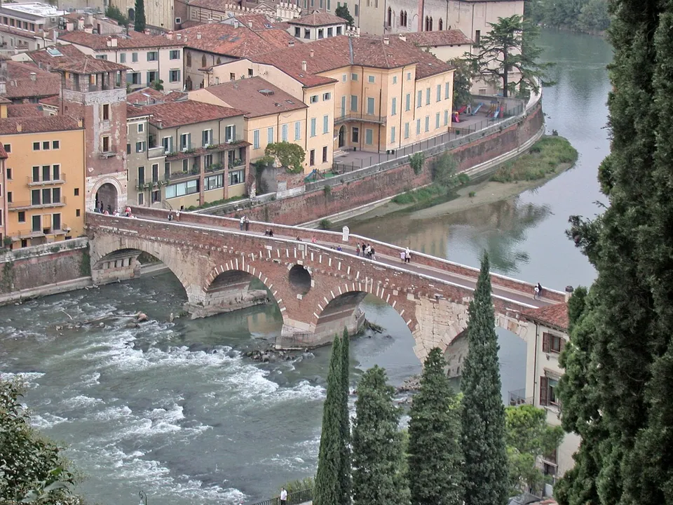 Looking down from a hillside between dark cypress trees on an arched bridge crossing a rushing river, with people walking across it, old houses on the far bank and a riverside road curving away beyond.