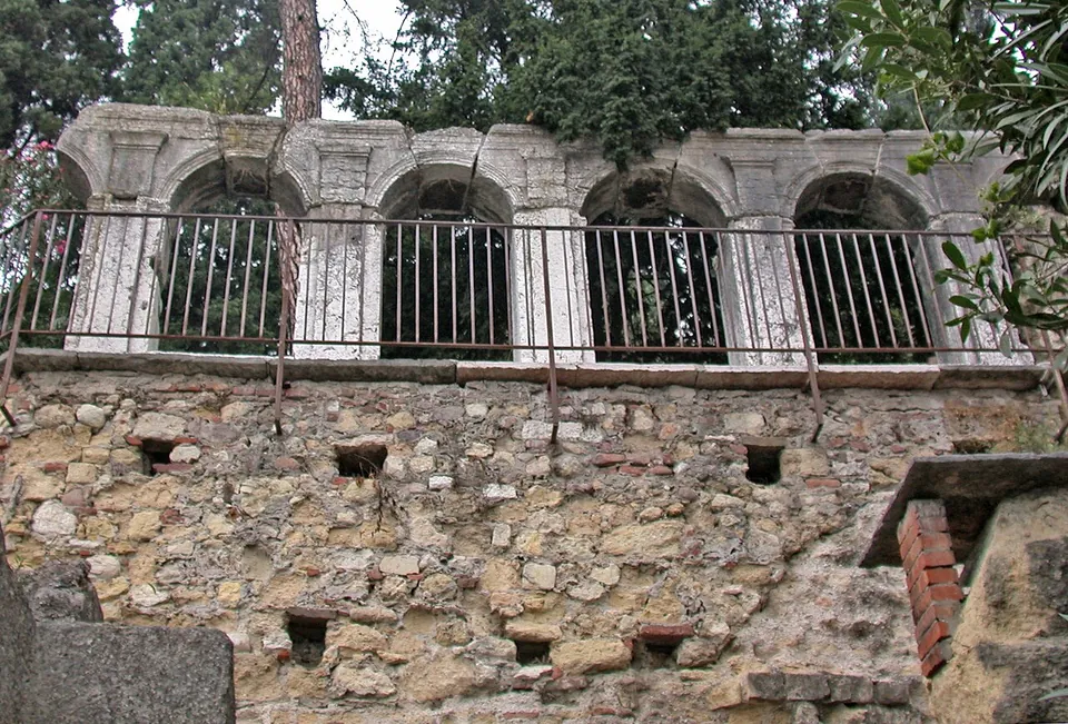 A row of round arches on slender stone piers standing along the edge of a terrace behind an iron railing, above a wall of rough stone and brick, with trees behind.