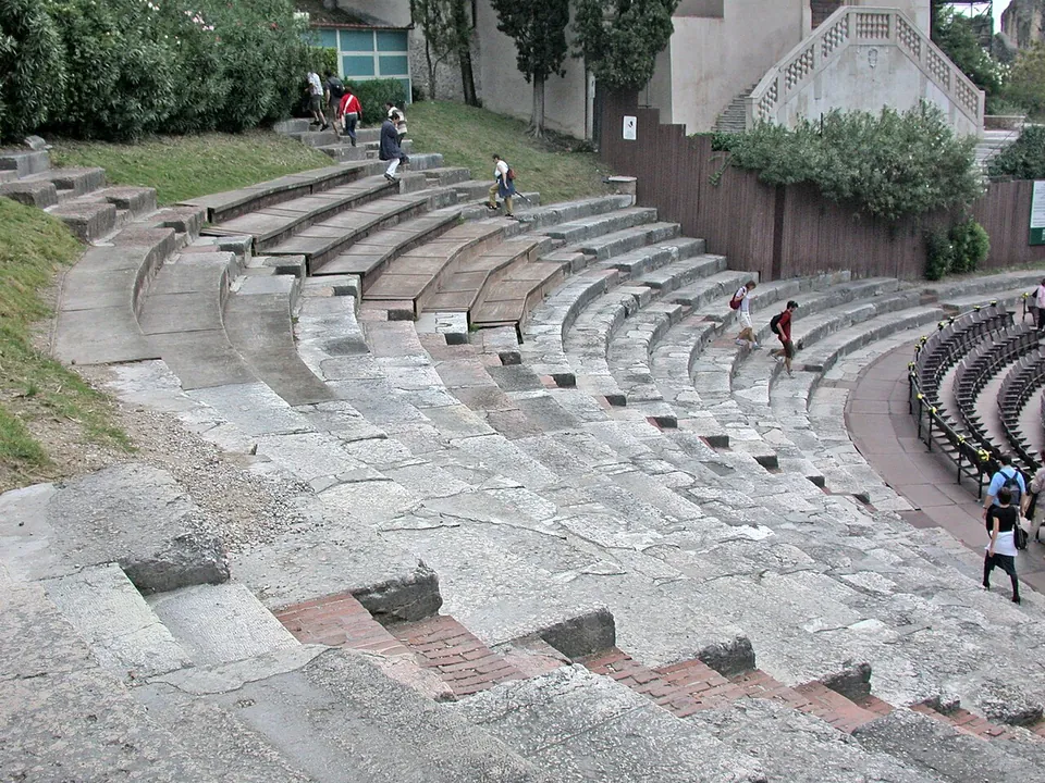 Curving tiers of worn grey stone seats climbing a grassy slope, with visitors walking along them and a stretch of red brick set into the lowest rows.