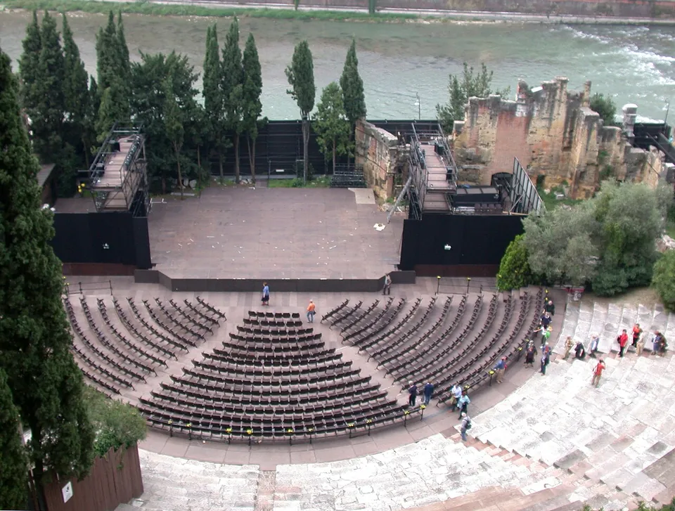 Looking down on a semicircle of stone seating with rows of modern seats set out on its lower part, facing a wide modern stage with lighting towers, ruined stone walls to one side and a fast river behind.