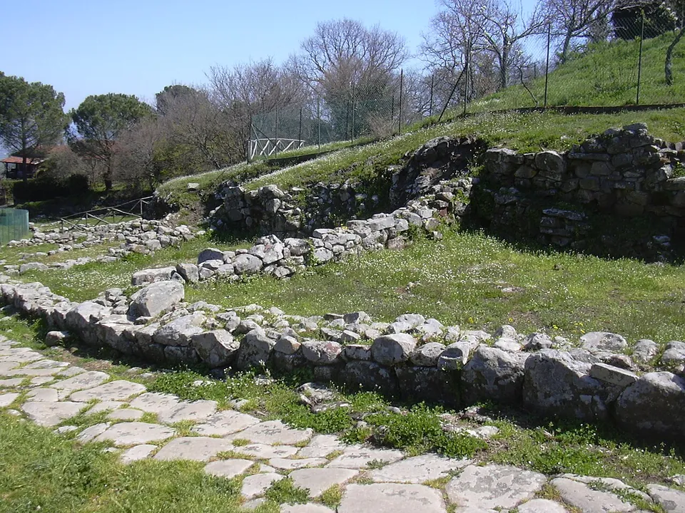A street paved with large flat stones runs past rows of low walls of rough grey stone, one to three courses high, set into a grassy slope with bare trees and a fence above.
