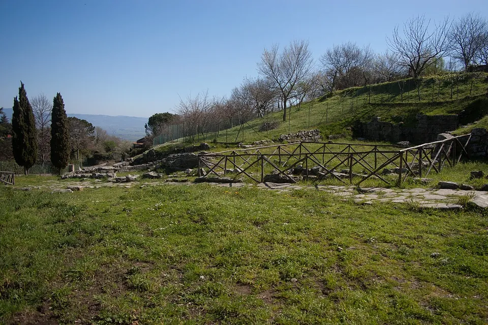 A grassy terrace with a wooden fence running along low excavated walls, cypresses to one side and a wide plain falling away to hills in the distance.