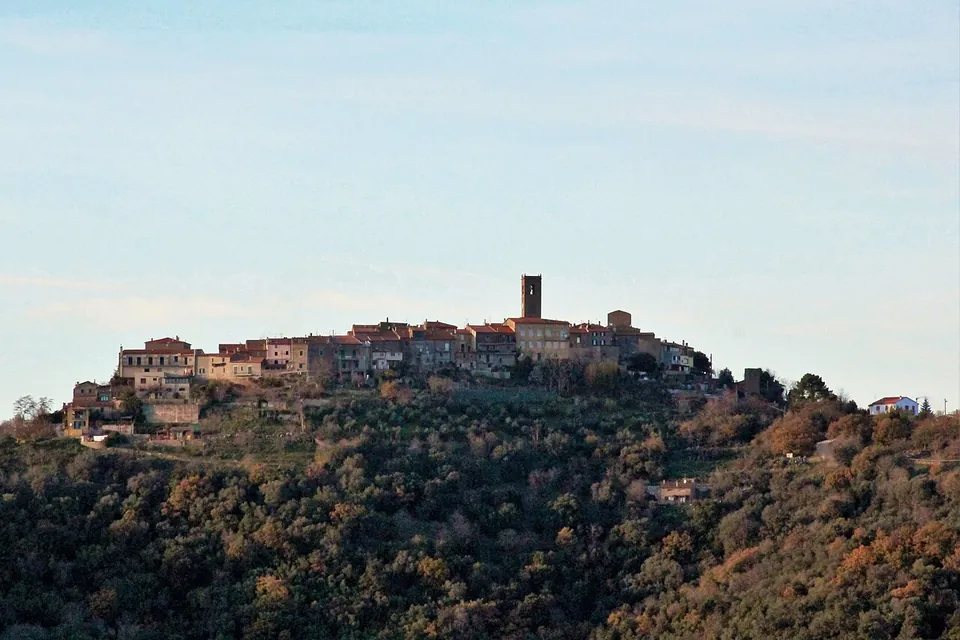 A small walled village of stone houses with a square tower strung along the crest of a wooded hill, under a pale evening sky.