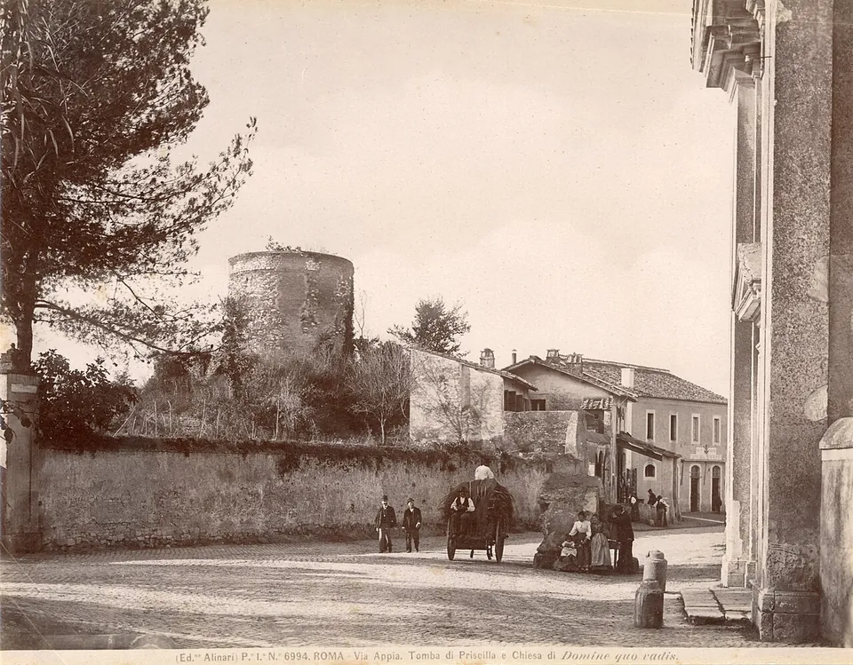 A sepia photograph of a road junction, with a cylindrical tower rising behind a long wall and low buildings, the corner of a church facade on the right, and a few figures and a horse-drawn cart in the road.