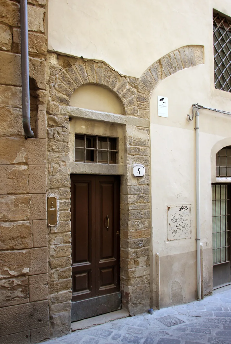 The front of a house in a narrow street, with a broad arch of rough brown stone blocks around a later doorway and fanlight, and the springing of a second stone arch beside it showing through the plaster.