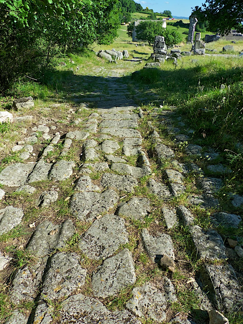 A road surface of large, irregular, worn paving stones running away between grassy verges, with scattered ruins and a standing column further along.
