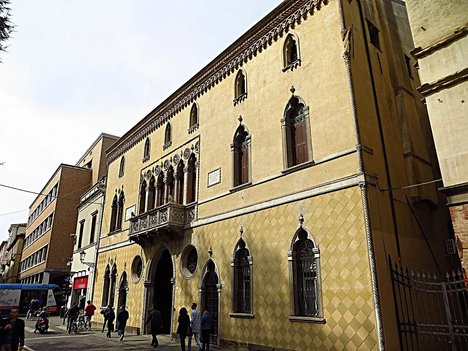 A street of old palazzi, the nearest with a ochre facade in a diamond pattern, pointed Gothic windows and a carved balcony, with pedestrians on the pavement and a bus further down.