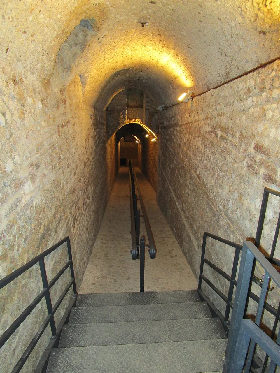 A narrow vaulted passage lit by lamps, running away into the distance, with modern steps and handrails leading down into it.