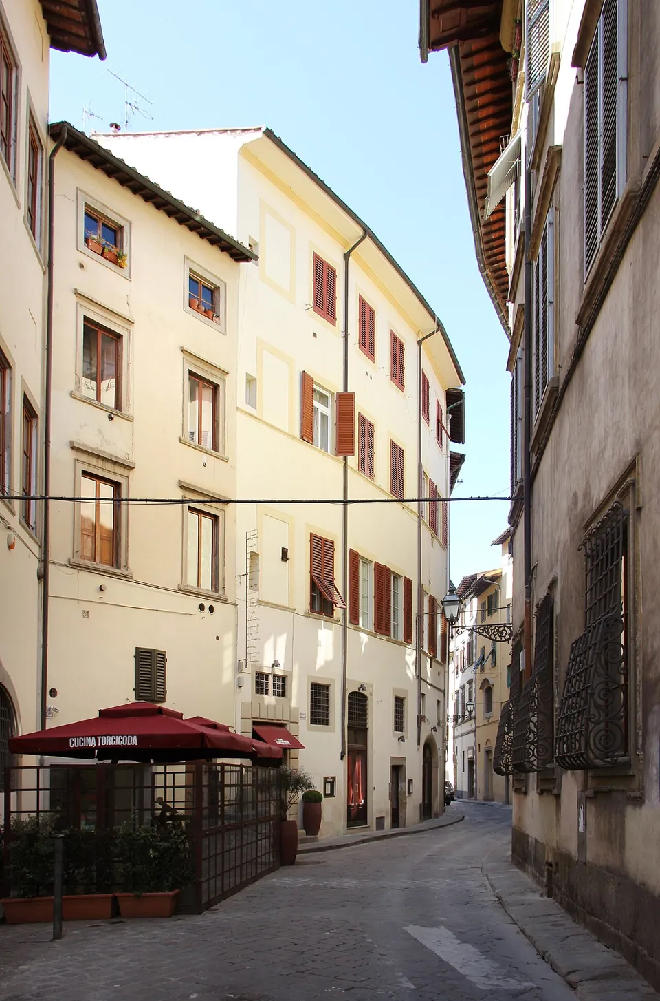 A narrow street of four-storey plastered houses that bends gently to the right as it runs away from the camera, with a café terrace under red umbrellas on the left.