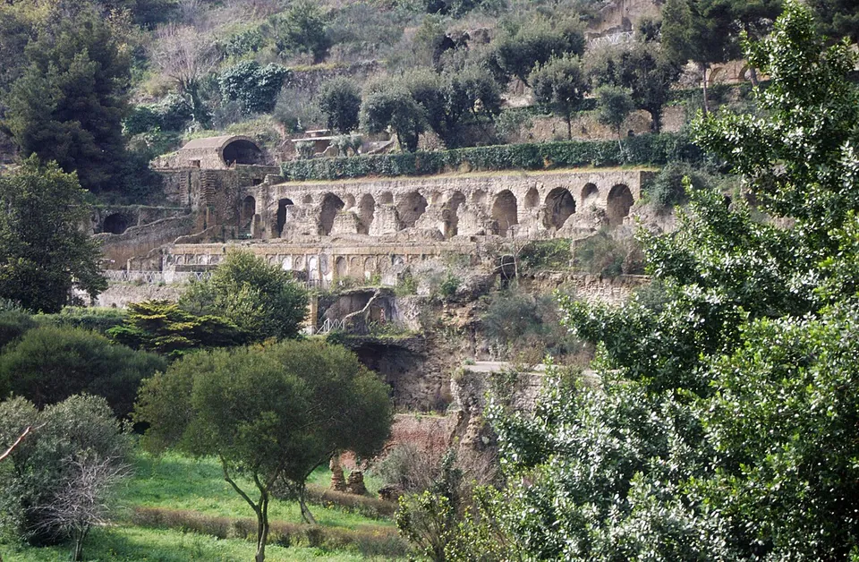 A hillside of ruins seen from below through trees, with a long row of arched openings along one terrace, walls and further terraces above it, and woodland at the top of the slope.