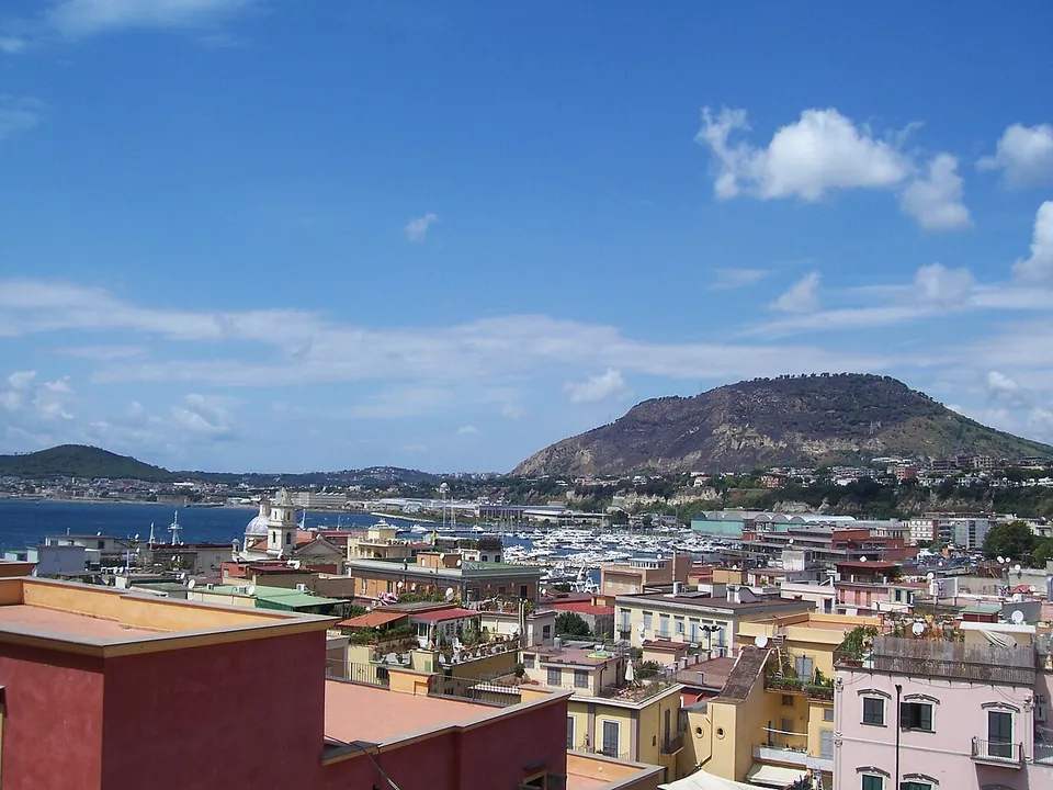 Pastel-coloured rooftops of the town running down to a harbour crowded with boats, with a broad green hill rising across the bay under a blue sky with scattered cloud.