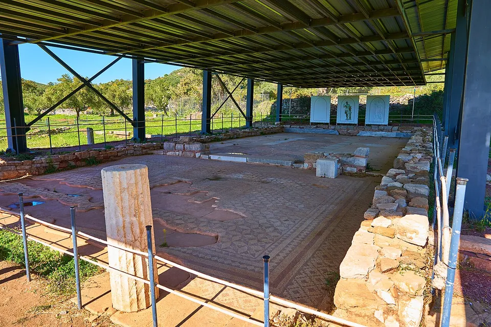 A worn mosaic floor and low stone walls under a modern metal roof on steel posts, fenced by a low rail, with three information panels at the far end.