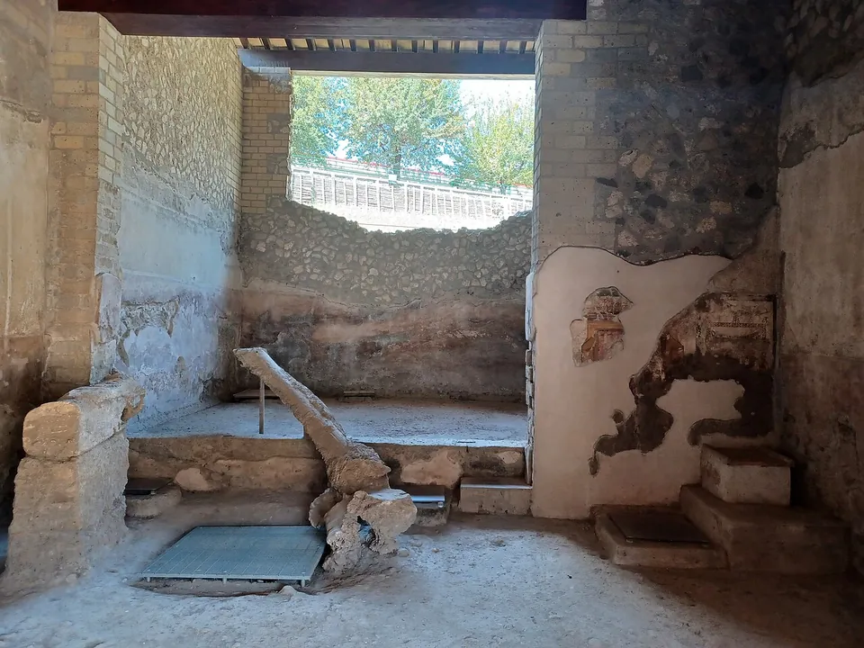 A room with rough stone and brick walls, patches of old plaster, a raised platform beneath a wide opening onto trees, and a long timber beam resting on stone blocks in the foreground.