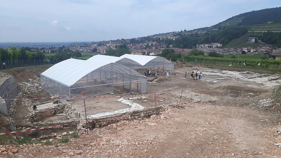 A broad excavation of bare earth and low stone walls, with white plastic tunnel shelters over part of it and a group of people standing among the trenches; rows of vines, a village and wooded hills rise behind.
