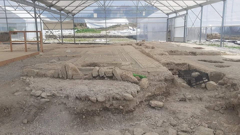 Inside a transparent temporary shelter on a metal frame, low rubble walls and a patch of mosaic floor lie in the excavated ground, with a wooden platform and railing at the left edge.