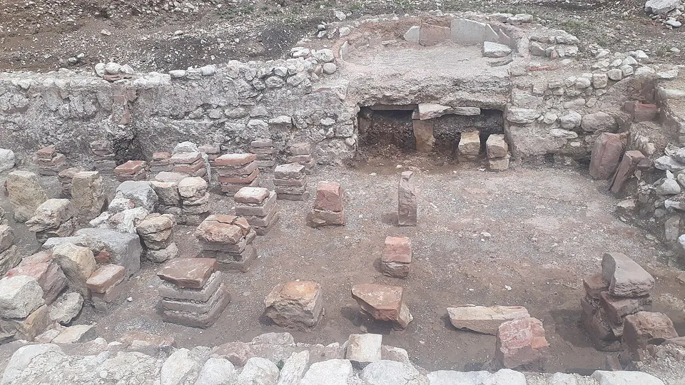 An excavated room enclosed by low walls of rough stone, its floor dotted with short stacks of square bricks and stone blocks, with an opening at the base of the back wall.