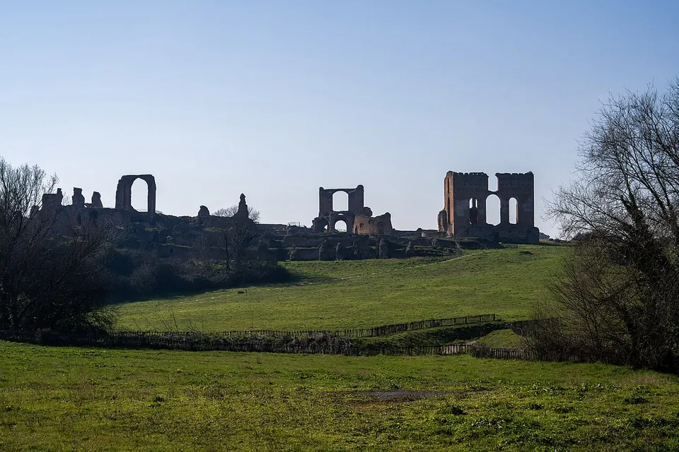 Across a wide green meadow and a low wooden fence, a long line of ruined brick walls stands on a rise, with tall arched openings and a block-like pair of towers at the right, bare trees at both edges and a clear sky.