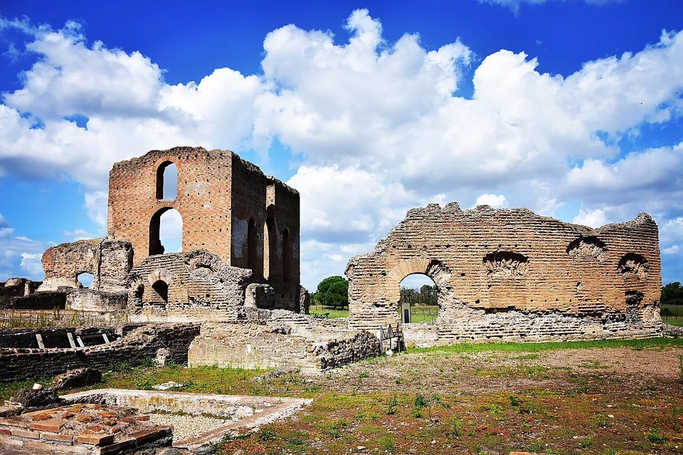 A tall block of brick wall with two large arched openings rises beside a long ruined brick wall with an arched doorway and a row of shell-like niches, above excavated floors and low walls in the foreground, under a sky full of white clouds.