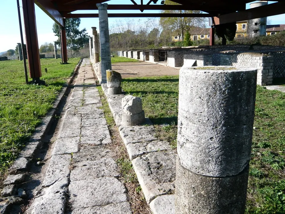 A paved walk runs between a row of stone columns and low rubble walls, under a modern timber roof on steel posts, with grass on either side and a building in the distance.