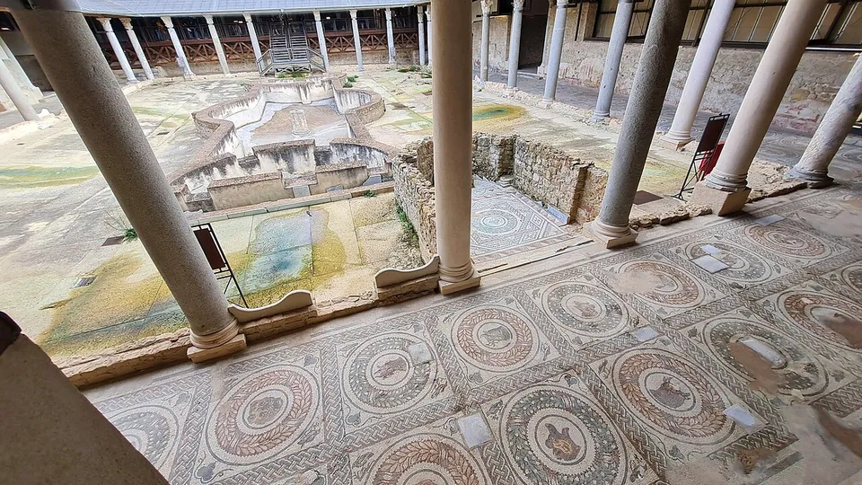 Looking out from a colonnade across a courtyard garden with a long stone fountain basin at its centre, the far side lined with columns under a modern roof; the portico floor in the foreground is a mosaic of round laurel wreaths, each framing an animal's head.