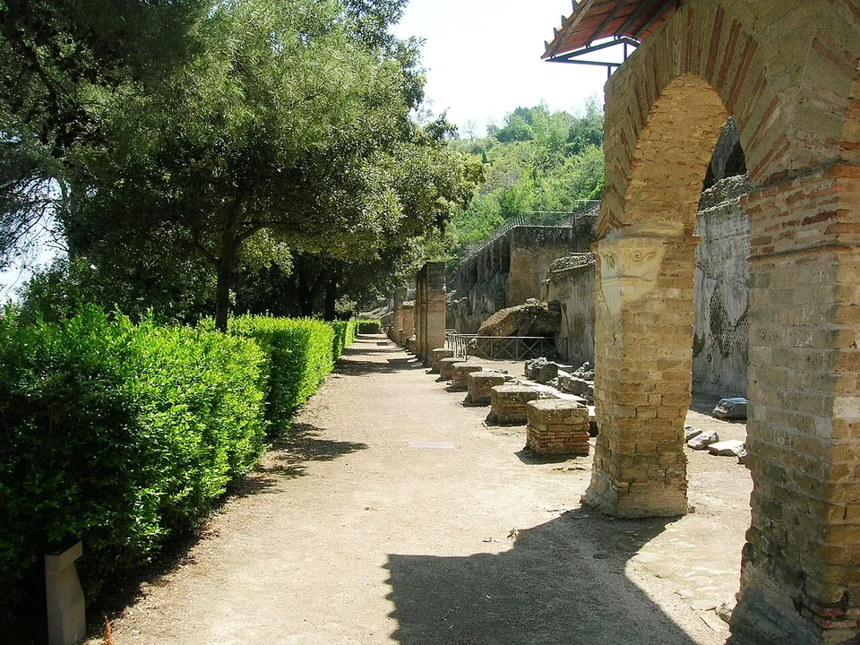 A gravel path running between a clipped hedge and a line of brick arches and pillar bases, under trees, with ruined walls further along.