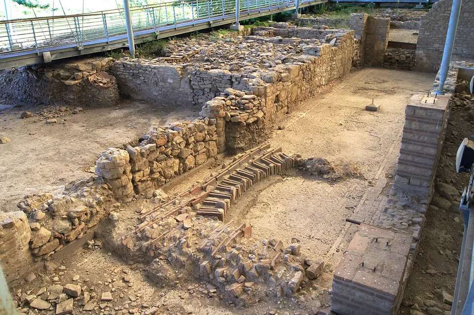 Excavated rooms of the villa seen from above, low walls of rough stone and brick around bare earth floors, a squared brick pier in the foreground and a raised metal walkway with railings running along the far side under the roof.